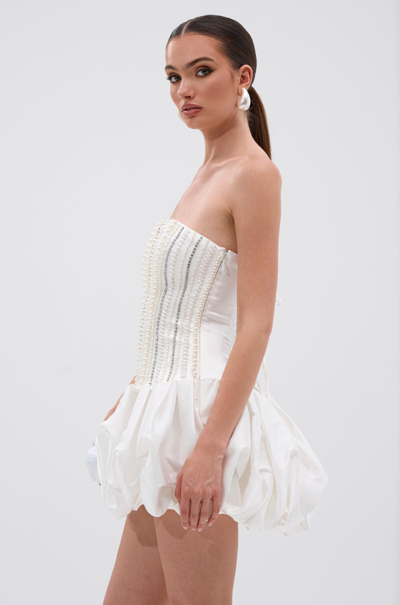 Model wearing the EXTRA! EXTRA! RHINESTONE MINI DRESS—a strapless white mini dress featuring a rhinestone corset-style lace-up bodice with pearl accents and a ruffled skirt. She has slicked-back hair in a low ponytail and large white hoop earrings.