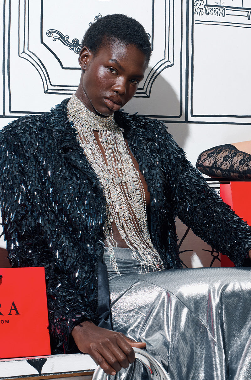 A model exudes luxurious style, wearing a shimmering black sequin jacket, the EXPENSIVE TASTE STATEMENT NECKLACE in silver fringe, and metallic silver pants. She poses confidently against a backdrop of illustrated architectural elements with a red box partially visible.