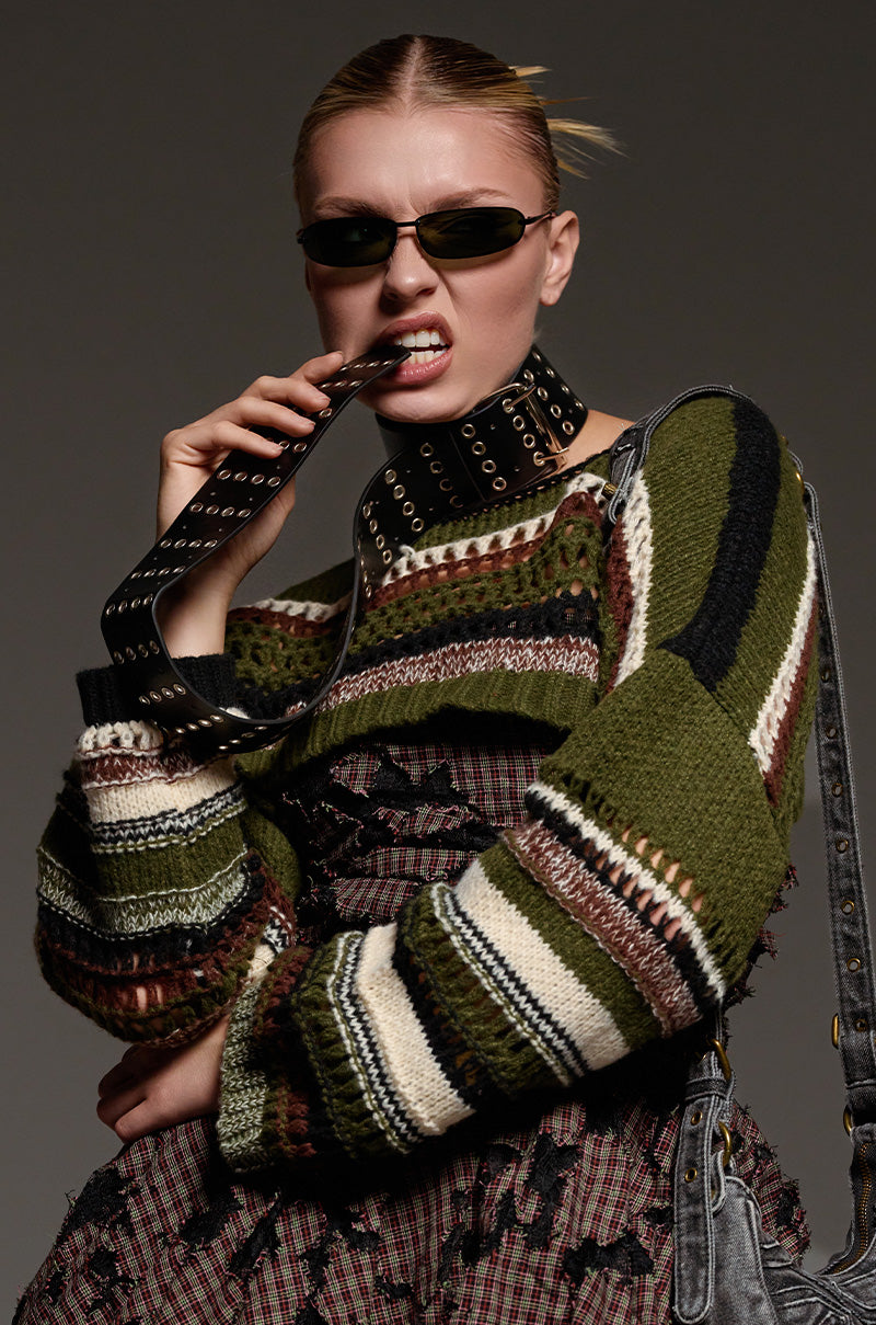 Wearing tinted sunglasses and a vibrant knit sweater adorned with stripes in black, green, and white, an individual creates a striking look by biting into a studded belt featuring a metallic silver buckle. Emphasizing their daring sense of fashion with a faux leather handbag draped over one shoulder, they set new style benchmarks with the High Standards Belt Necklace.