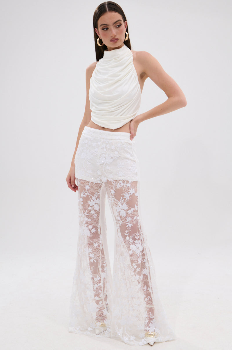 A woman poses against a plain white background wearing the UP NEXT LACE PANT IN WHITE, a high-waisted floral lace pant with sheer panels, paired with a sleeveless white draped top, gold hoop earrings, and straight hair.