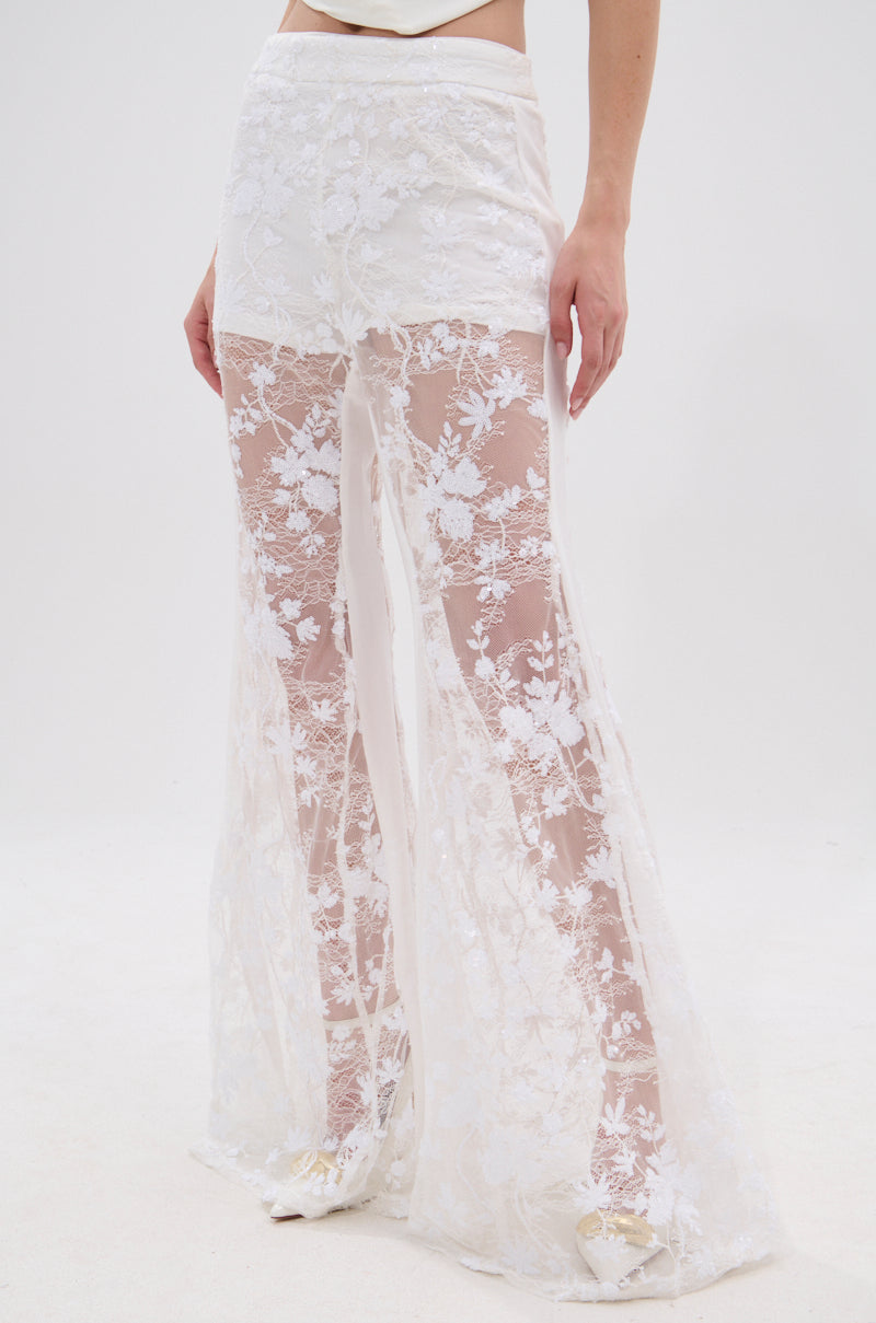 Wearing the UP NEXT LACE PANT IN WHITE, which features high-waisted, sheer floral lace and flared legs, a person stands against a plain white background with only their lower torso and legs visible.