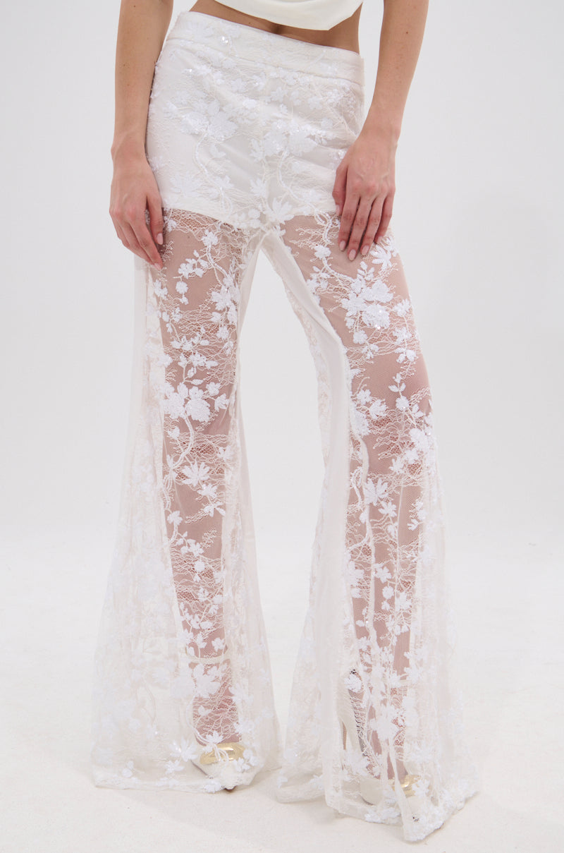 Wearing the UP NEXT LACE PANT IN WHITE, a person showcases sheer, high-waisted floral lace flare pants with delicate embroidery, revealing their legs as they rest their hands on their thighs against a plain white background.