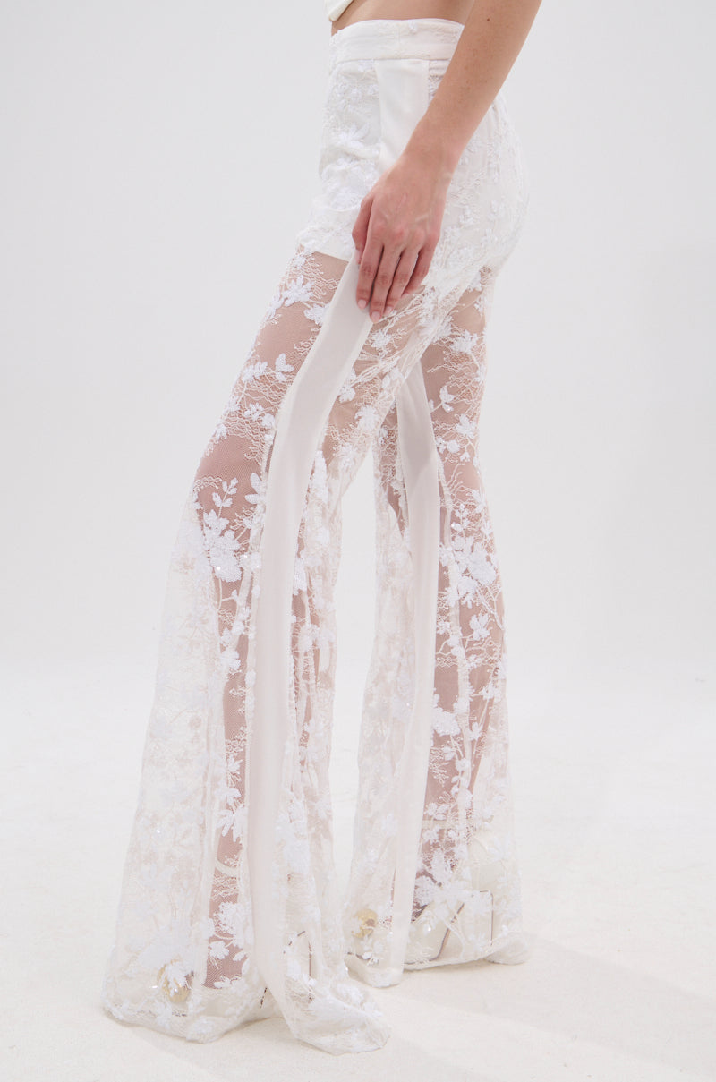 A person wearing the UP NEXT LACE PANT IN WHITE, featuring high-waisted, sheer floral lace with flared legs and delicate patterns, stands against a plain white background.