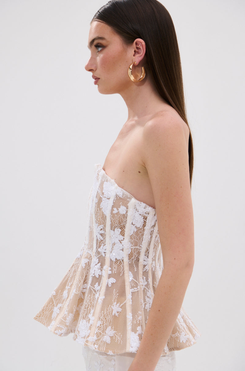 A woman with straight brown hair wears gold hoop earrings and the UP NEXT LACE CORSET TOP IN WHITE, featuring white floral lace and a peplum flare, standing in profile against a plain light background.