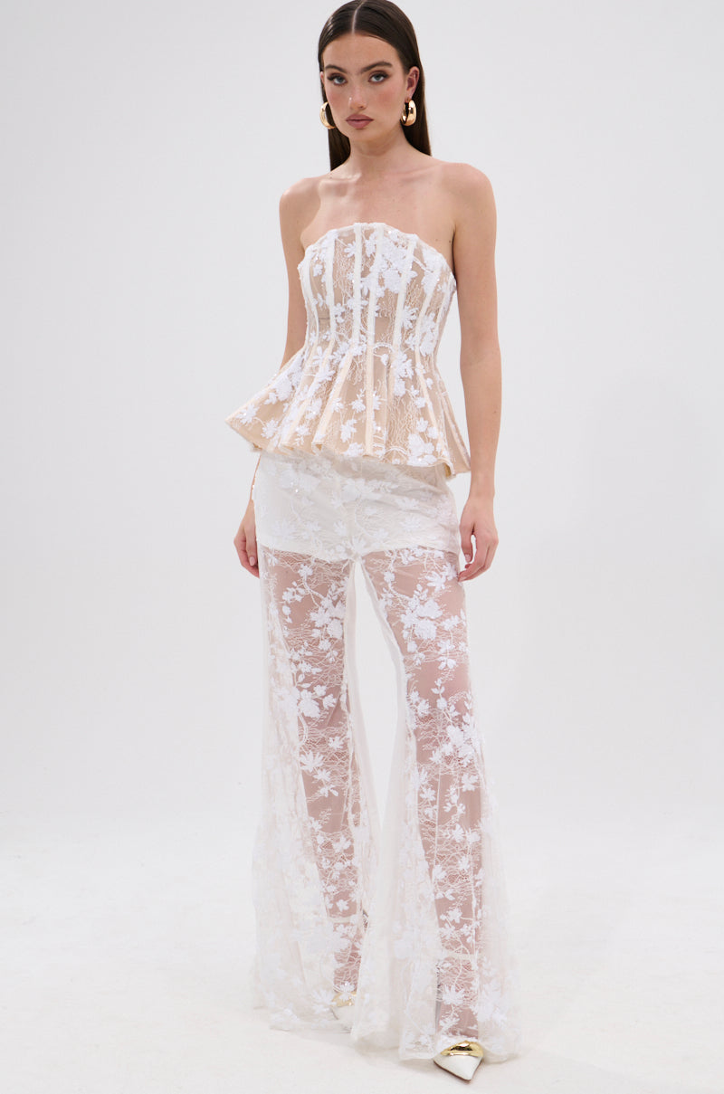 A woman models the UP NEXT LACE CORSET TOP IN WHITE, a strapless floral lace peplum flare corset, paired with matching sheer flared pants, gold statement earrings, and pointed shoes against a white background.