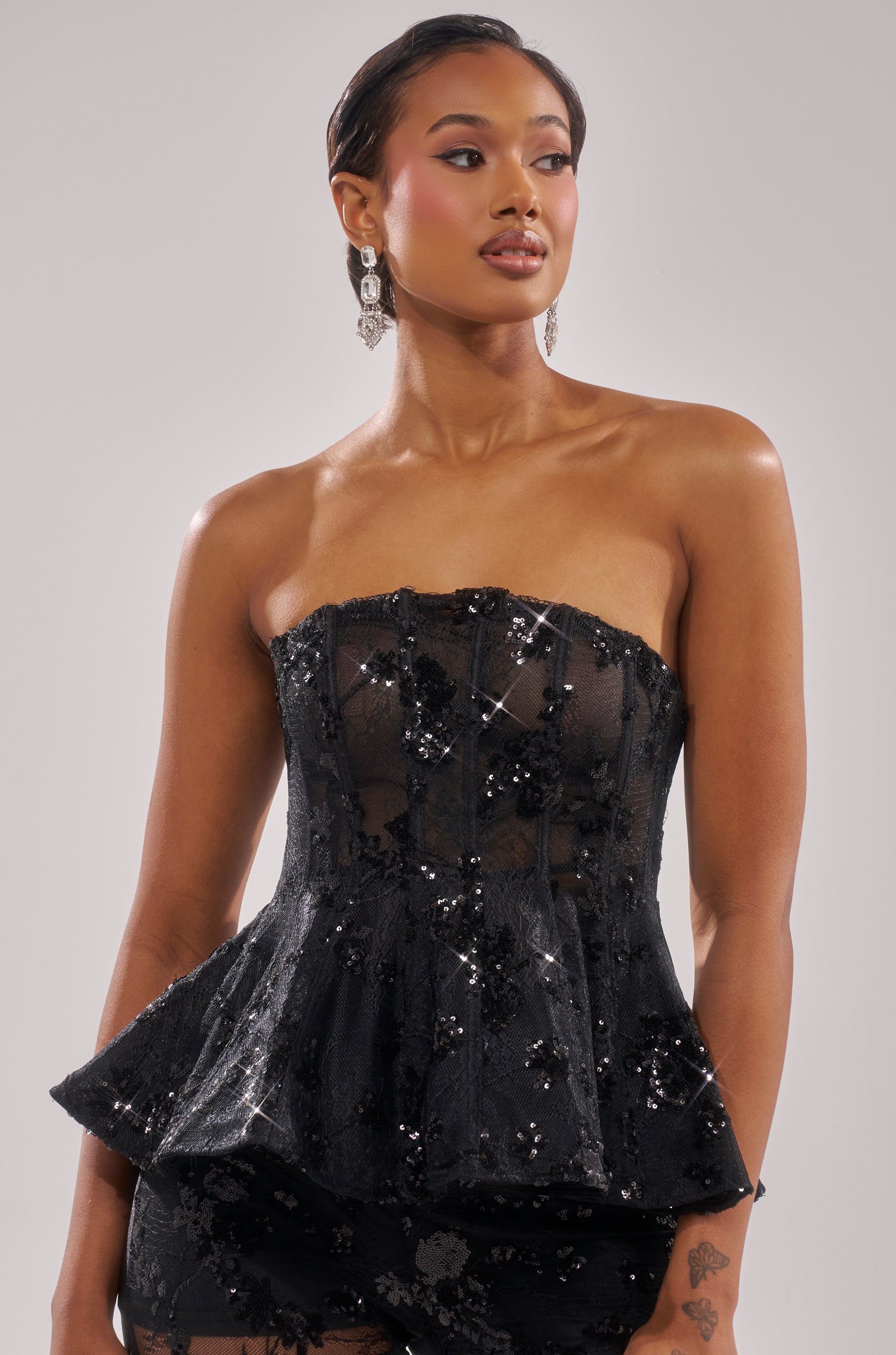 A woman poses in the UP NEXT LACE CORSET TOP, featuring a strapless black sequin peplum design. She has slicked-back hair, dangling earrings, and gazes to the side against a plain background with a neutral expression.