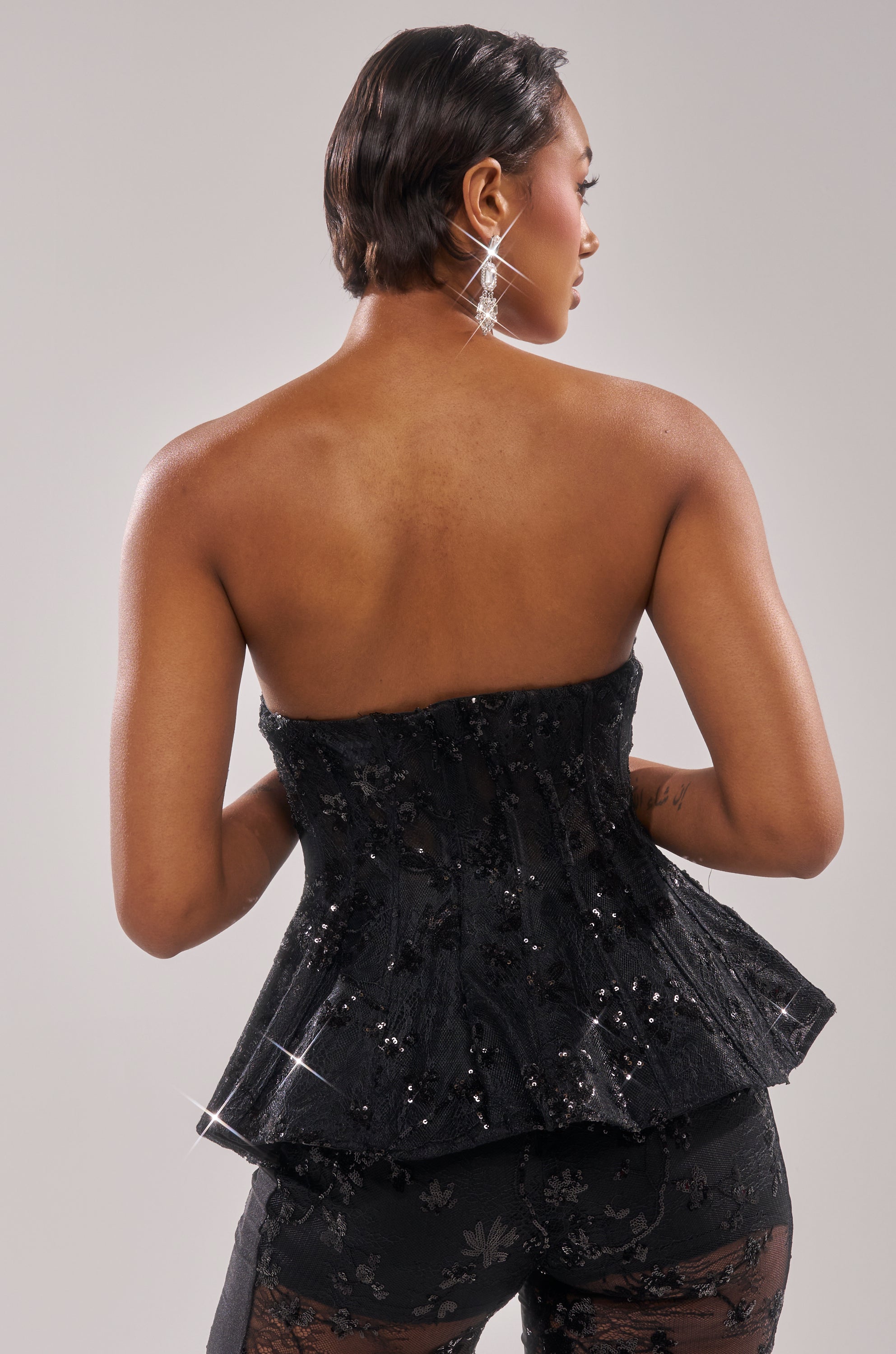 A woman with short hair poses with her back to the camera, wearing the UP NEXT LACE CORSET TOP and matching black lace pants, accessorized with dangling earrings against a plain light background.
