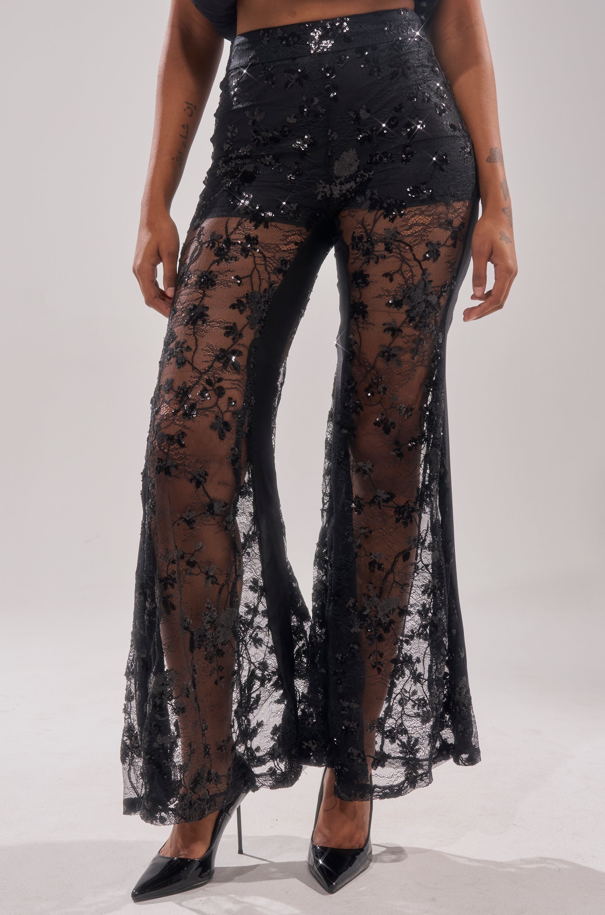 A woman wears the UP NEXT LACE PANT, featuring high-rise, flared legs with floral patterns and sequin details. Sheer from mid-thigh down, the look is finished with black pointed-toe heels.