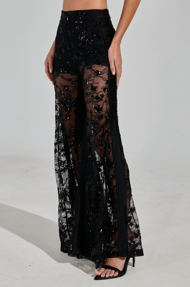 A woman wears stunning UP NEXT LACE PANT from the AKIRA Label, featuring floral embroidery and sequins. These chic pants pair perfectly with black high-heeled sandals against a plain, light gray background.