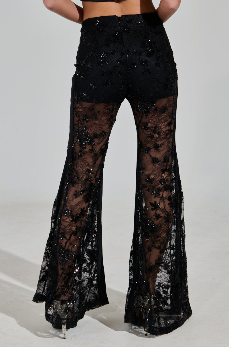 The person is wearing UP NEXT LACE PANT by AKIRA Label, featuring black floral lace with sequin details and a solid waistband, standing out beautifully against the light-colored floor.