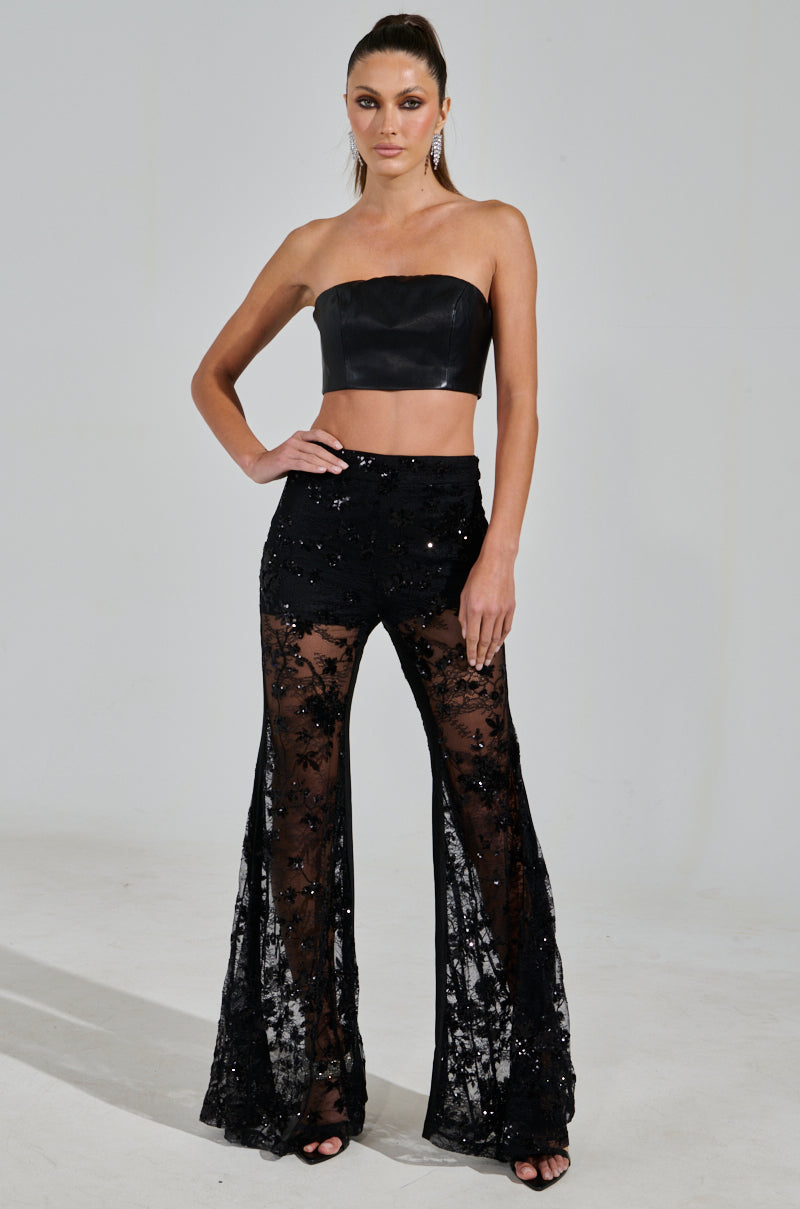 A model confidently poses against a plain white background wearing AKIRA Label's UP NEXT LACE PANT featuring sheer black lace and sequins, paired with a strapless black leather crop top. She completes the look with open-toe black heels, earrings, and a sleek ponytail.