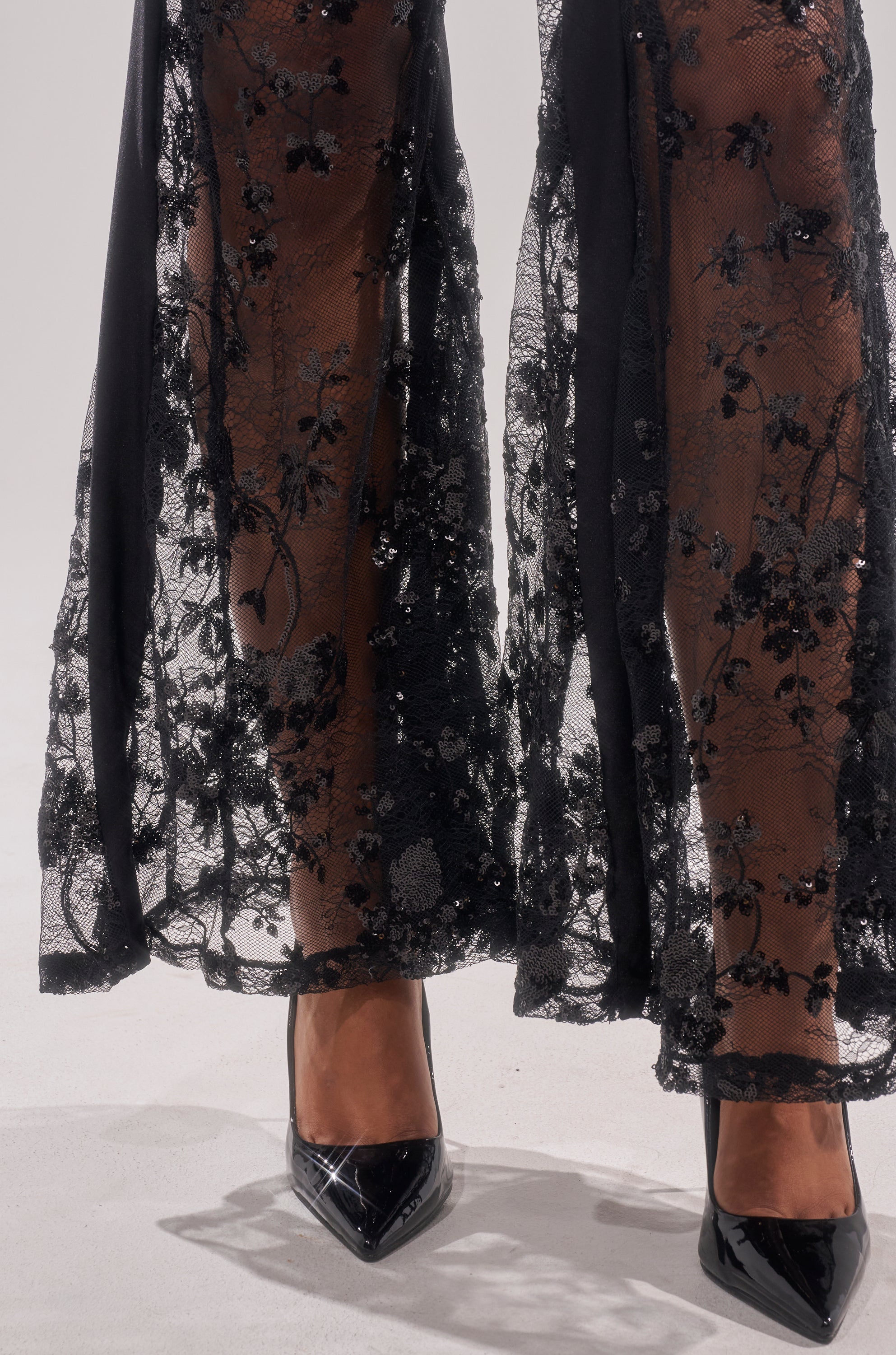 A close-up of a person wearing the UP NEXT LACE PANT, a high-rise sheer black lace pant with floral patterns, paired with black patent leather pointed-toe heels on a white surface.