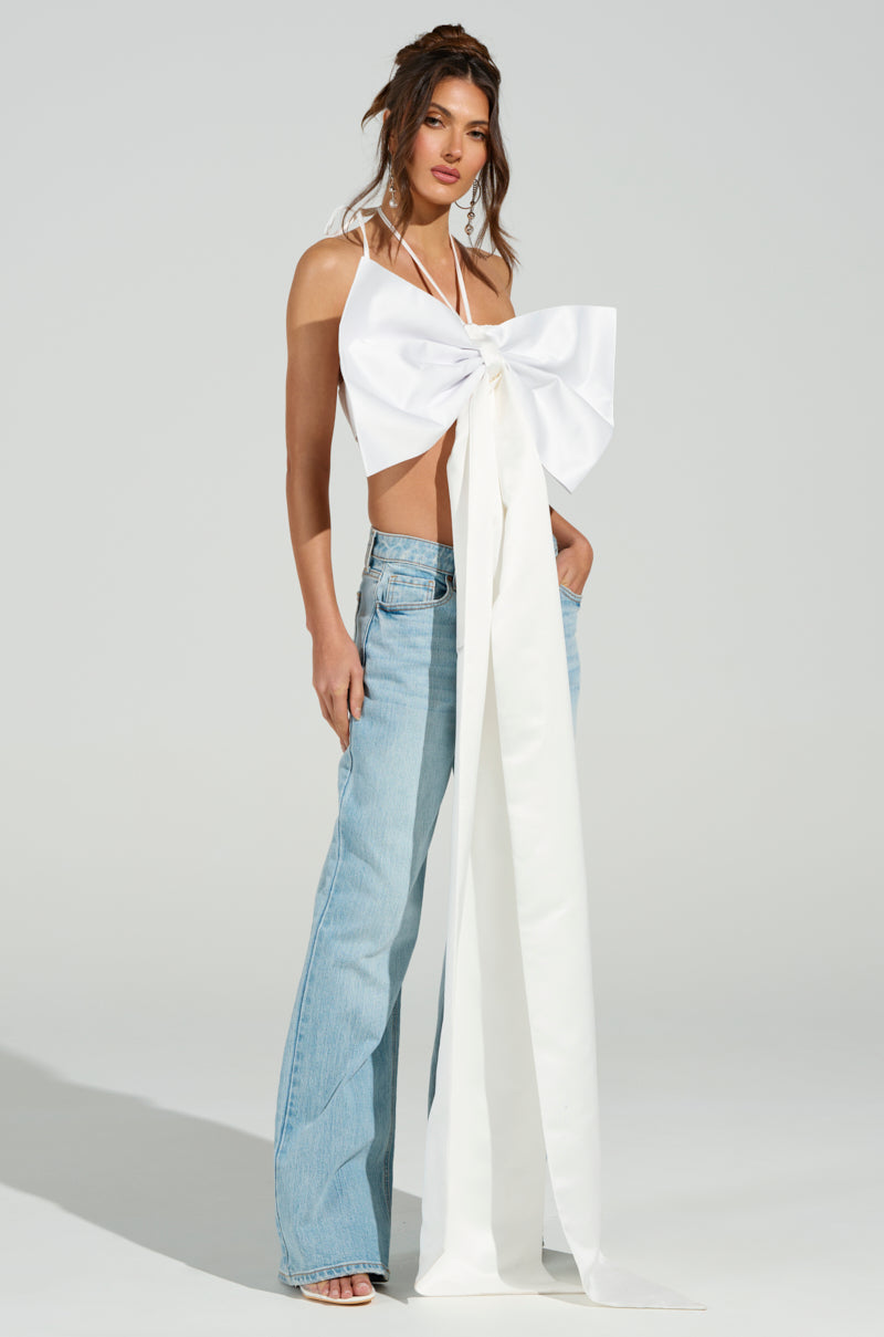 A woman models the "WRAP ME UP IN A BOW TOP," a satin halter top styled as a large white bow, paired with light blue flared jeans and white heels. The metallic back zipper adds an edge as she poses with one hand in her pocket against a plain gray background. 0