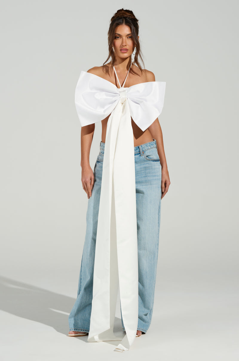 A woman poses against a plain backdrop in light blue wide-leg jeans and the WRAP ME UP IN A BOW TOP, featuring long trailing ends. The satin fabric of the top adds subtle shine, while her loosely waved hair complements her elegant appearance. 4