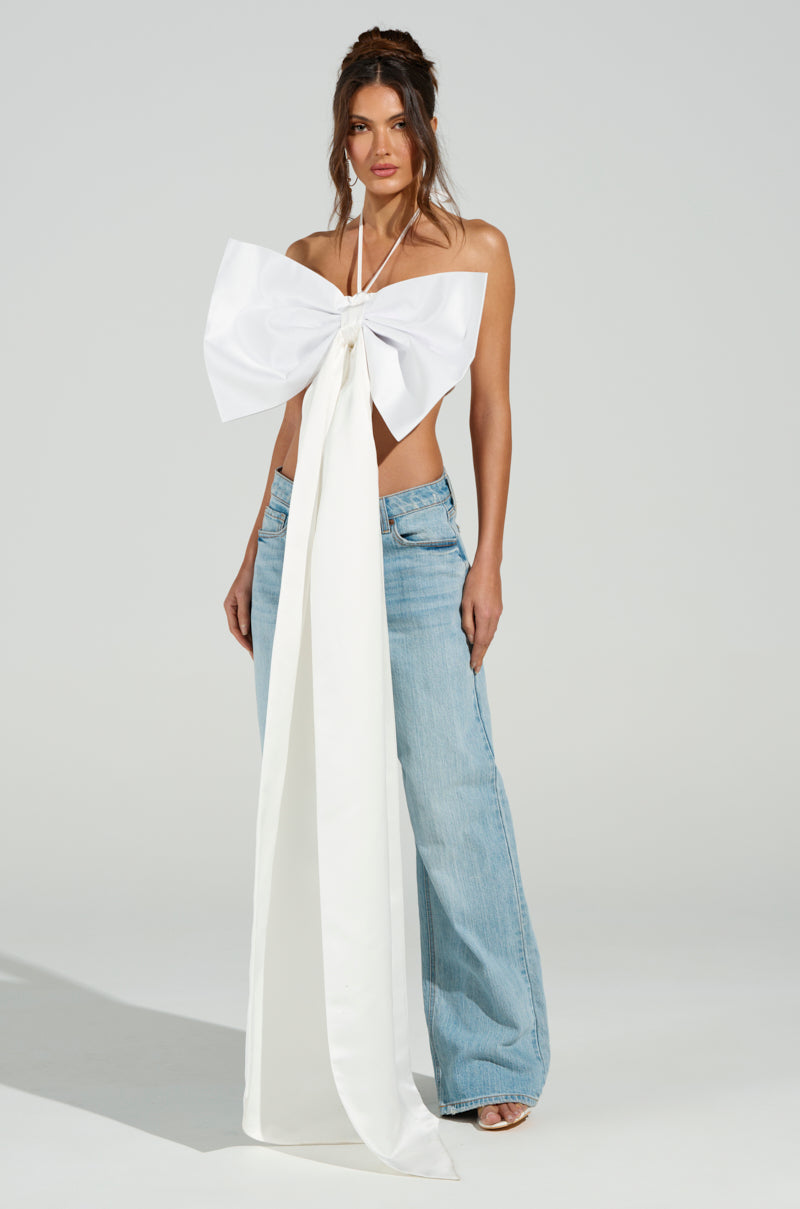 A woman models the "WRAP ME UP IN A BOW TOP," showcasing its large white satin bow with long tails, paired with blue wide-leg jeans. Her casual updo and the metallic back zipper add an edge, while the plain background emphasizes her chic ensemble. 5