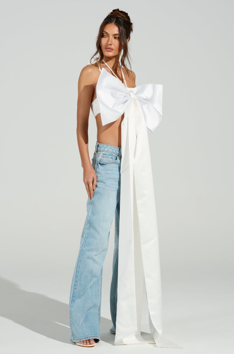 A woman in the WRAP ME UP IN A BOW TOP with satin fabric, long ribbons, and a metallic back zipper is paired with light blue wide-leg jeans and white heels. Her long brown hair styled in an updo against a plain light gray background. 6