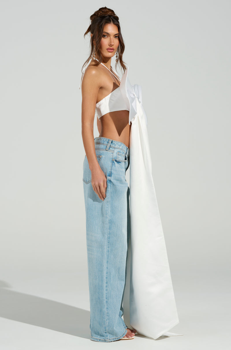 A woman poses against a plain backdrop, wearing the WRAP ME UP IN A BOW TOP in white satin featuring a metallic back zipper and pairing it with light blue wide-leg jeans. Her hair is styled up with loose strands framing her face as she gazes towards the camera. 7