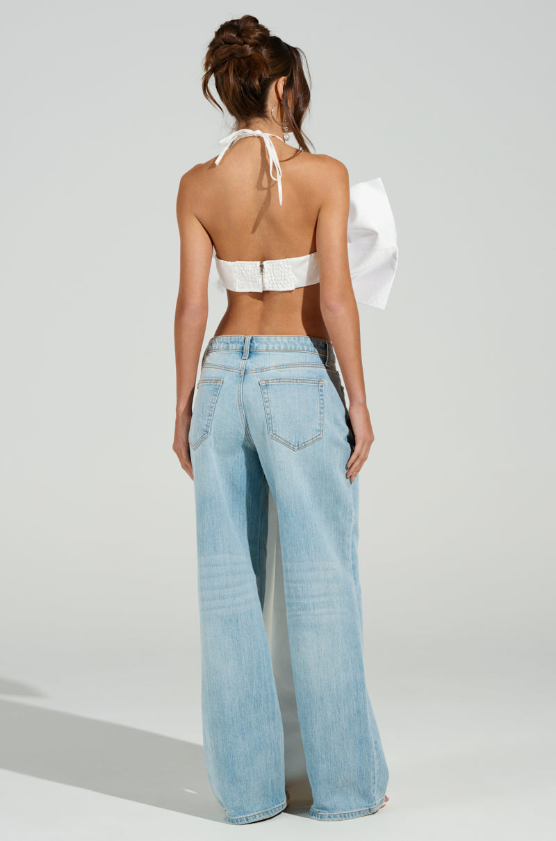 With their long hair cascading down, the person faces away in a chic outfit—a WRAP ME UP IN A BOW TOP with a metallic back zipper and wide-legged light blue jeans. The satin halter top adds elegance, while the neutral background enhances the casual look. 8