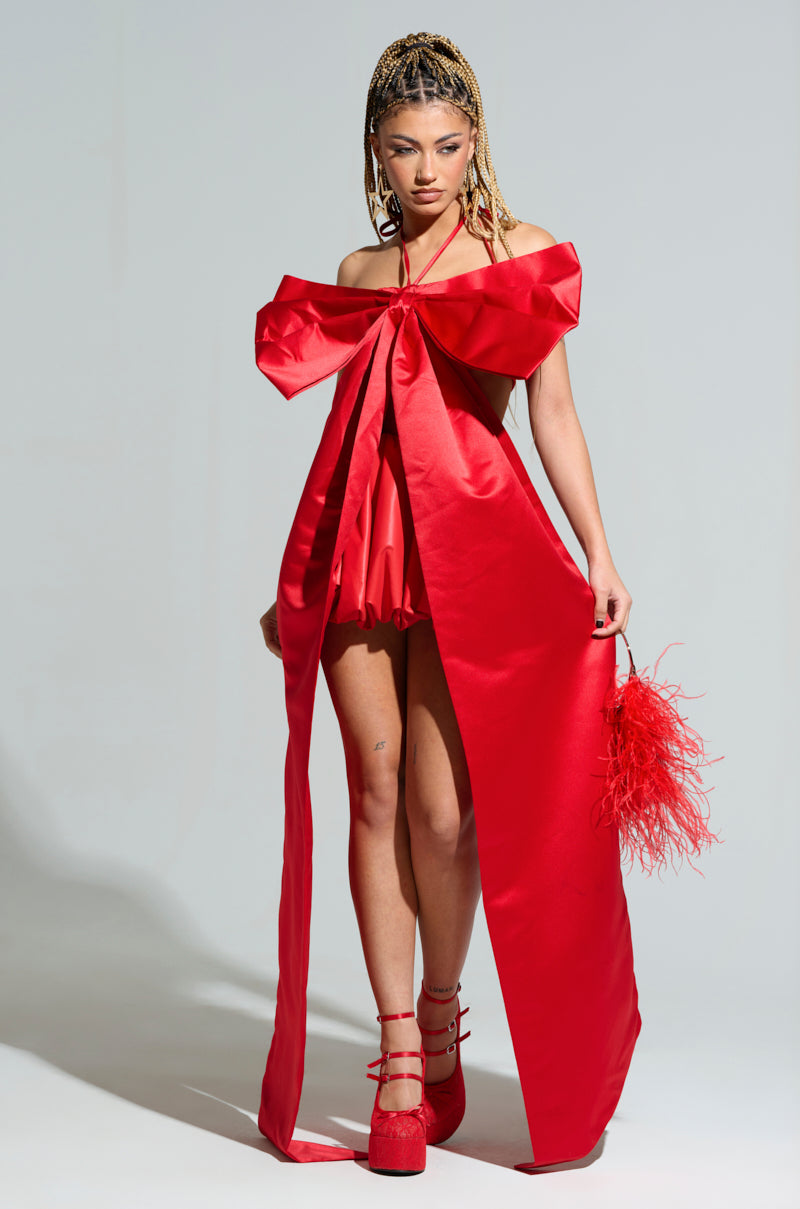 A person in a striking red dress reminiscent of the WRAP ME UP IN A BOW TOP IN RED, featuring flowing ribbons on the sides. They hold a matching red feathered handbag and wear red platform shoes, all set against a plain light gray background.