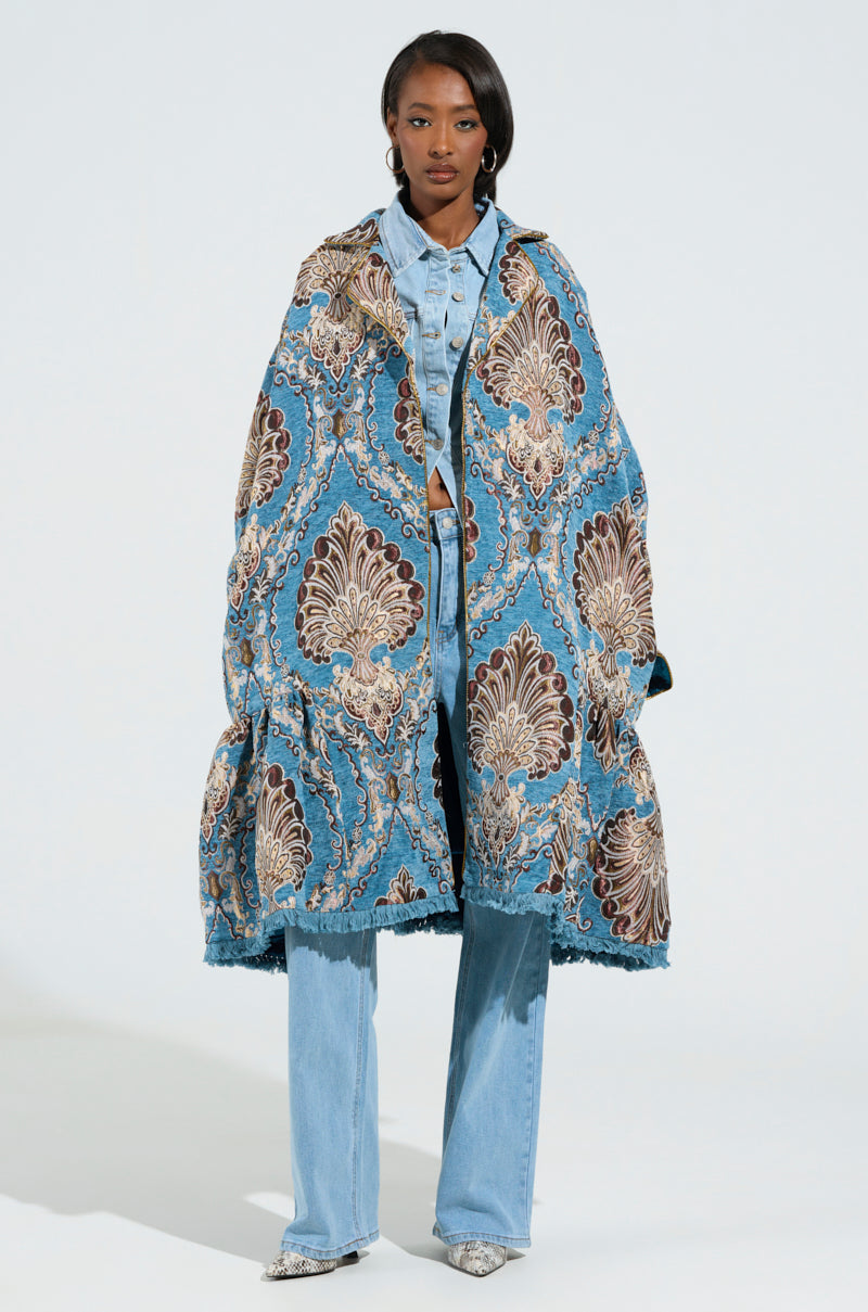 A person wearing a long, patterned trench coat with the Kensington Tapestry Trench design in blue and brown is layered over a denim shirt and light blue jeans, standing against a plain background. Their pointed shoes perfectly match the colors of the coat. 0