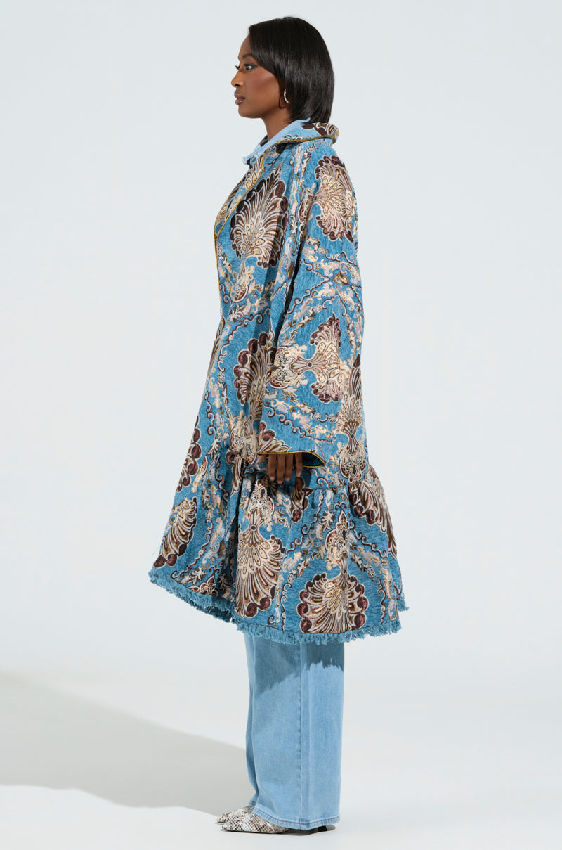 A person in profile is wearing a Kensington Tapestry Trench, featuring an oversized longline silhouette with an intricate floral design. The trench coat is paired with light blue jeans and complements their straight black hair, all set against a simple, light background. 2