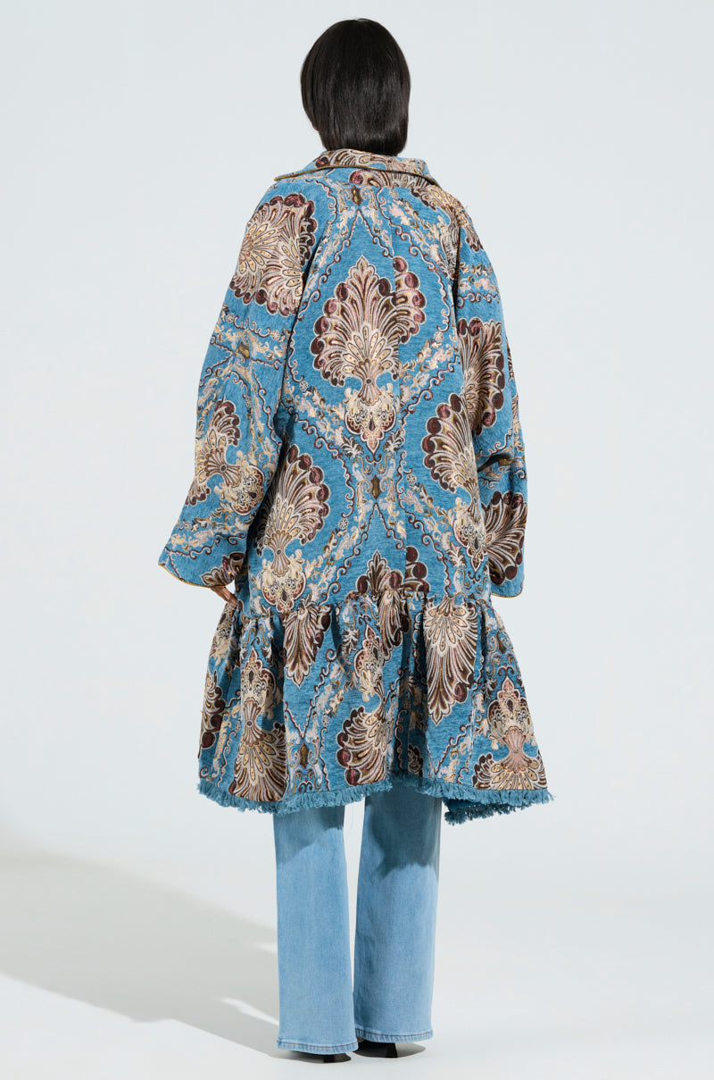 A person wearing the KENSINGTON TAPESTRY TRENCH, featuring wide sleeves and a ruffled hem, stands with their back turned, showcasing light blue jeans beneath. The oversized longline silhouette is highlighted by the vibrant blue and brown floral pattern set against a solid white backdrop. 3