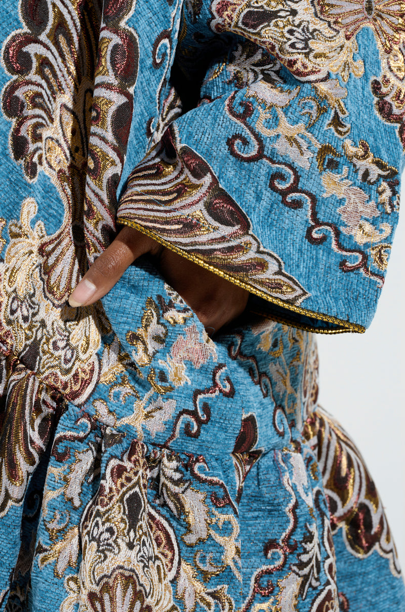 A close-up shot showcases someone wearing the KENSINGTON TAPESTRY TRENCH in a striking blue and gold palette, featuring an oversized longline silhouette adorned with intricate floral designs. The luxurious fabric, with its metallic details and rich textures, is elegantly highlighted as the person gently rests their hand on it. 4