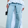 A person elegantly flaunts an asymmetrical high-low silhouette in wide-leg blue jeans and a black cropped top. They wear the RUNWAY BELT SKIRT draped over the pants and hold a small black handbag, adding a touch of high-fashion allure.