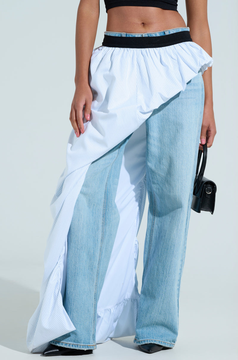 Someone is wearing the RUNWAY BELT SKIRT, a ruffled, asymmetrical high-low piece over denim jeans, with a black crop top. They carry a chic small black handbag against a plain light backdrop.