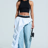 Wearing sunglasses and a black crop top, she pairs light blue wide-leg jeans with an asymmetrical white RUNWAY BELT SKIRT. A small black handbag completes her high-fashion look against a light gray background.