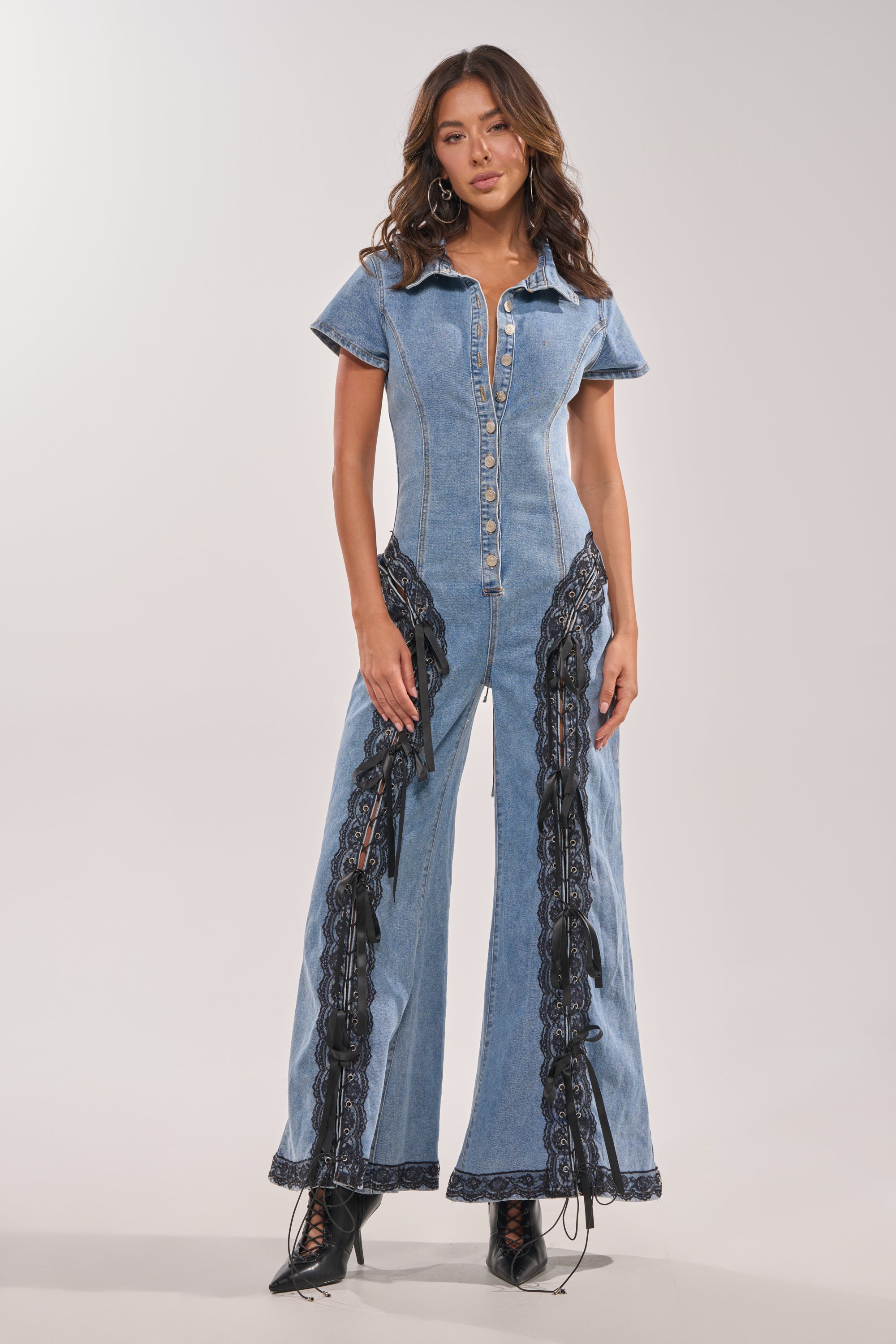 A woman models the DIXIE LACE DENIM JUMPSUIT, a short-sleeved, button-up piece with black lace sides and front, styled with black lace-up boots and loose wavy hair against a plain light background. 2