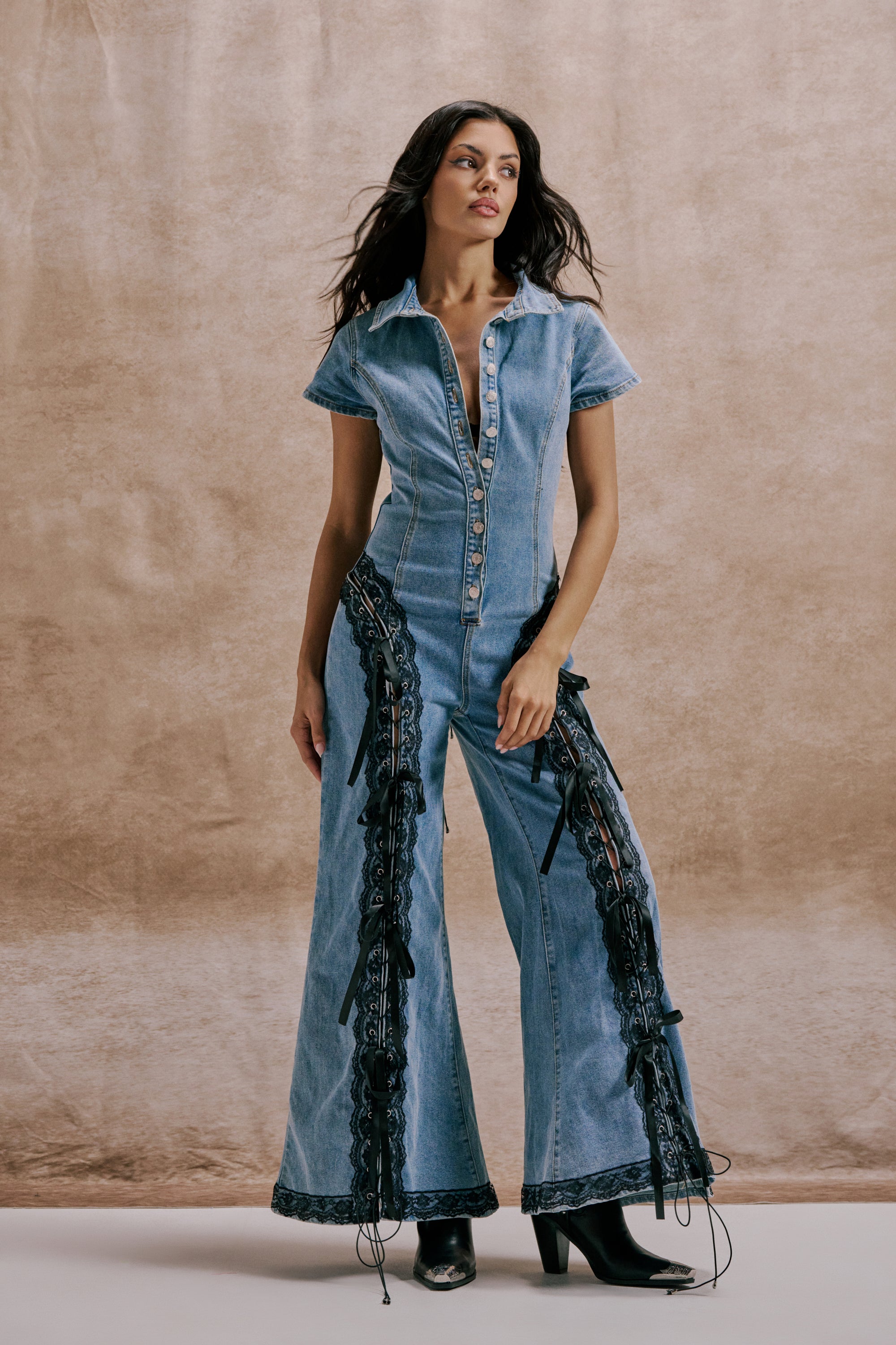 A woman poses against a beige backdrop, wearing the DIXIE LACE DENIM JUMPSUIT featuring short sleeves, flared legs, black lace accents, and paired with black heeled boots. 0
