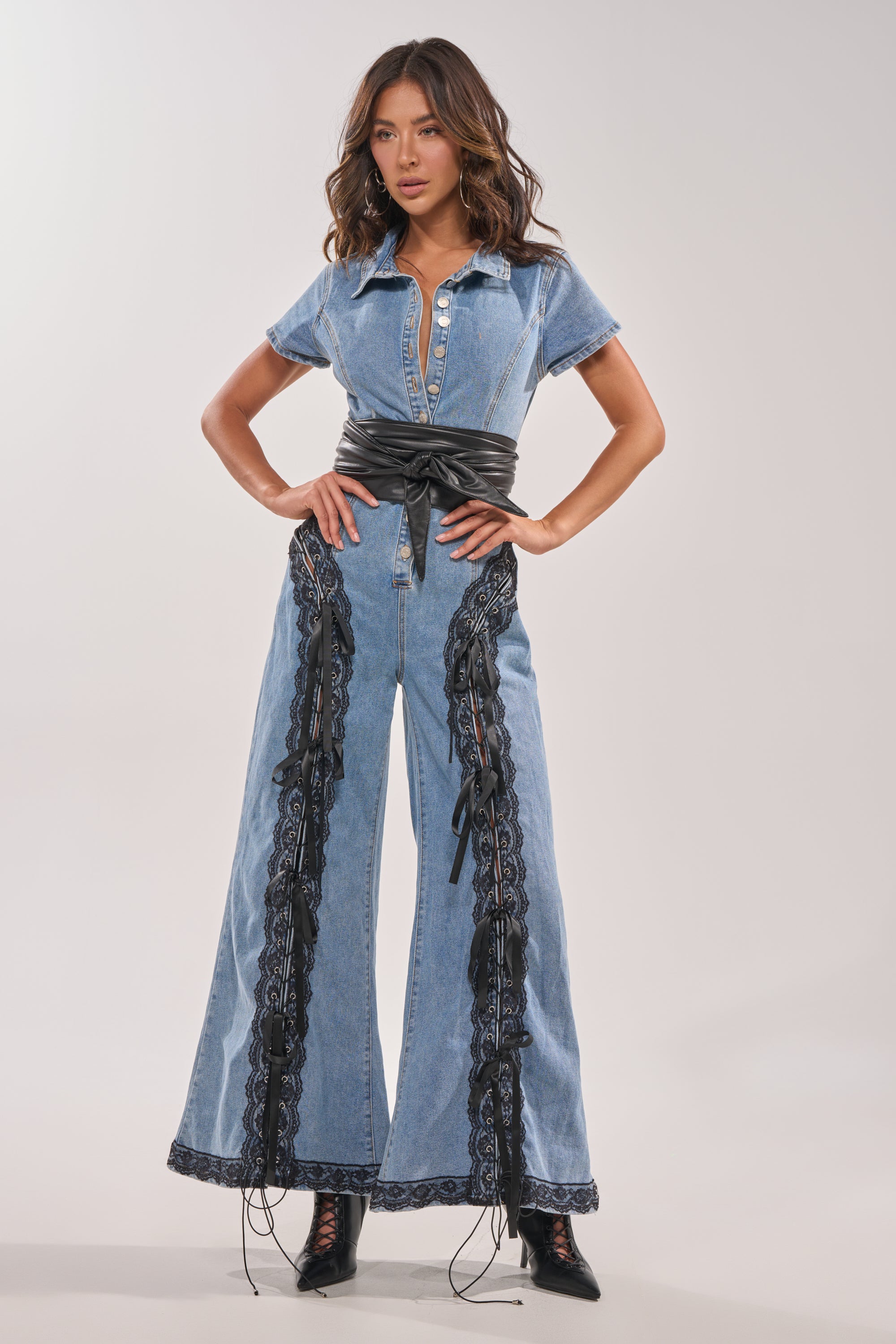 A woman wears the DIXIE LACE DENIM JUMPSUIT featuring short sleeves, a flared leg with black lace details, styled with a black wide belt and lace-up boots, against a plain light background. 3