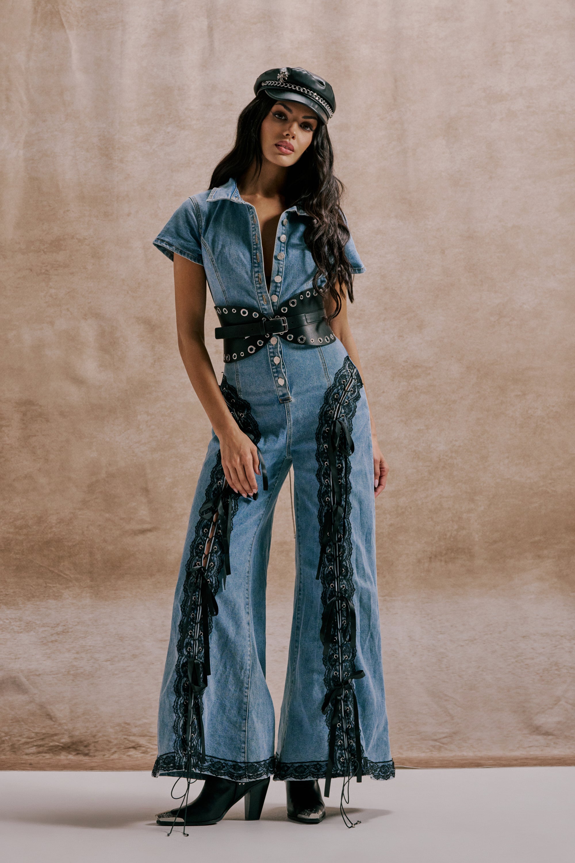 A woman poses before a beige backdrop in the DIXIE LACE DENIM JUMPSUIT, featuring black lace details, flared legs, a wide grommet belt, matching hat, and sleek black heeled boots. 1
