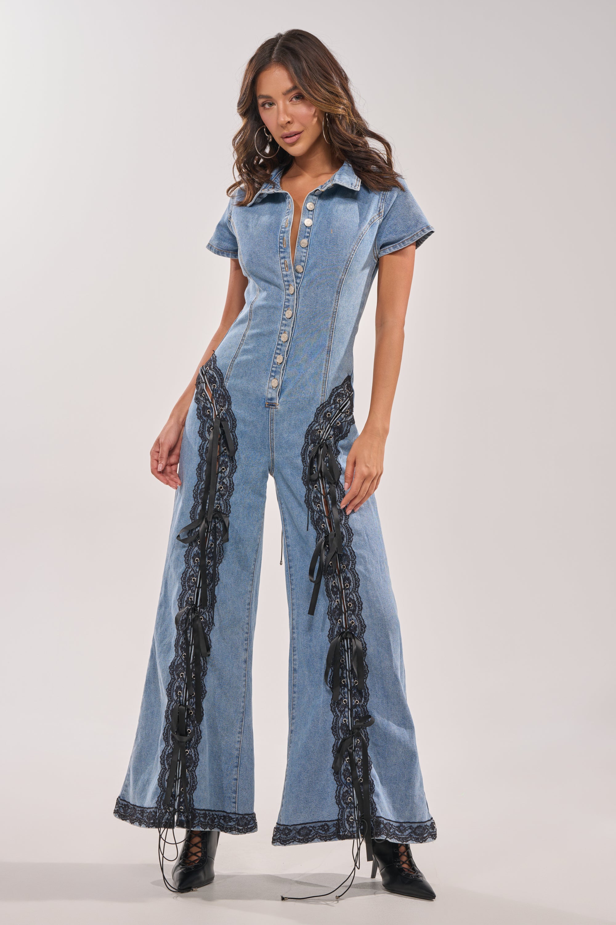 A woman confidently models the DIXIE LACE DENIM JUMPSUIT, featuring short sleeves, a button-up front, flared legs, and black lace panels with ties down the sides, standing against a plain light background. 4