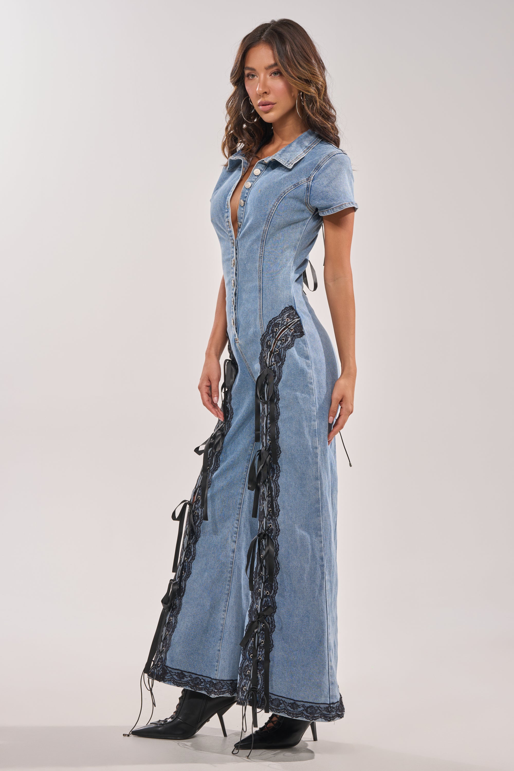 A woman models the DIXIE LACE DENIM JUMPSUIT, a long, fitted piece with black lace detailing on the sides and hem, short sleeves, and front buttons. She completes the look with pointed black boots against a light background. 6