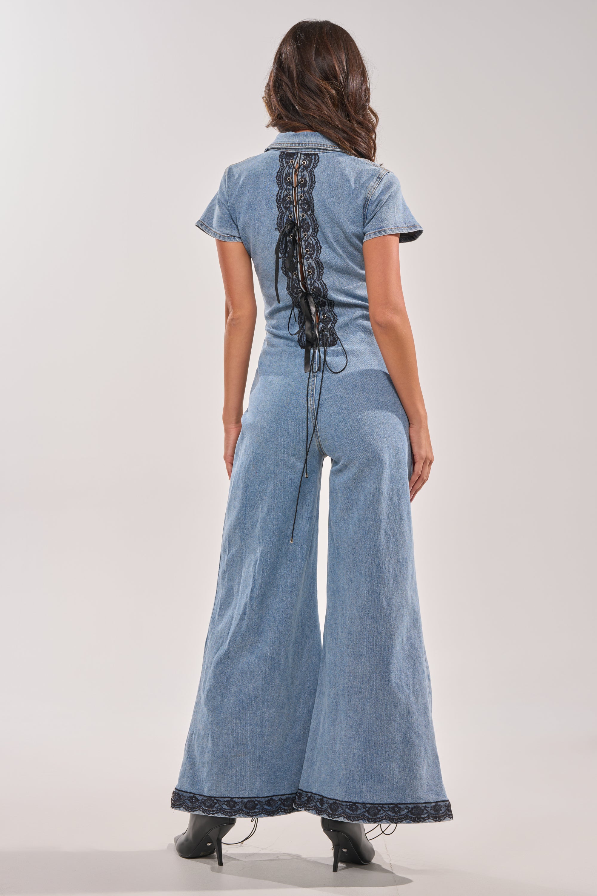 A woman faces away in the DIXIE LACE DENIM JUMPSUIT, featuring a light blue wash, flared legs, short sleeves, and black lace running down the back. She pairs it with black shoes and wears her wavy brown hair loose against a plain background. 7