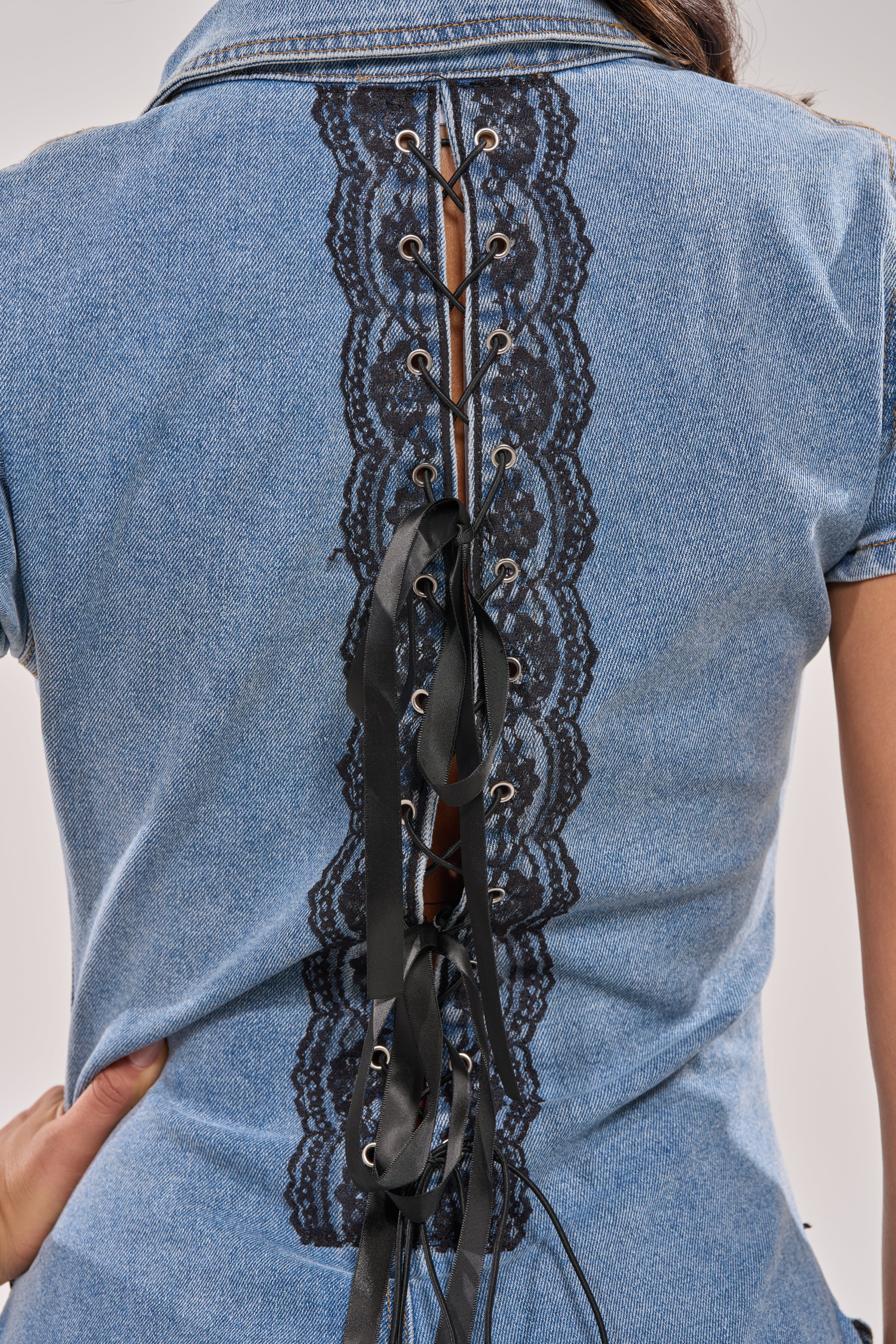 A close-up of a person wearing the DIXIE LACE DENIM JUMPSUIT, featuring black lace details, ribbon corset lacing up the back, and modern flared legs as their hand rests on their hip. 8