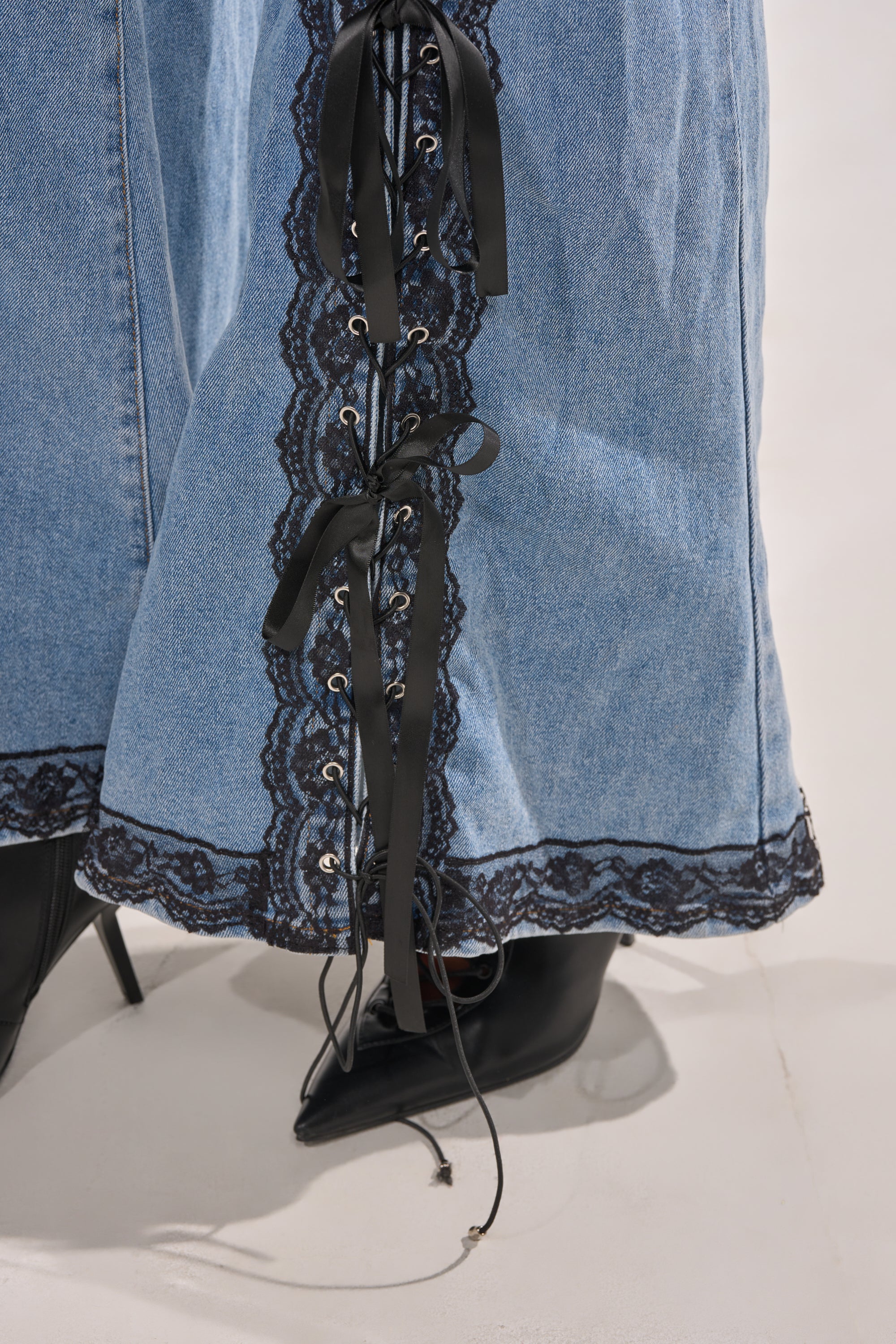 Close-up of the DIXIE LACE DENIM JUMPSUIT with black lace detailing and ribbon lacing on the side, styled with pointed black high-heeled boots. 9