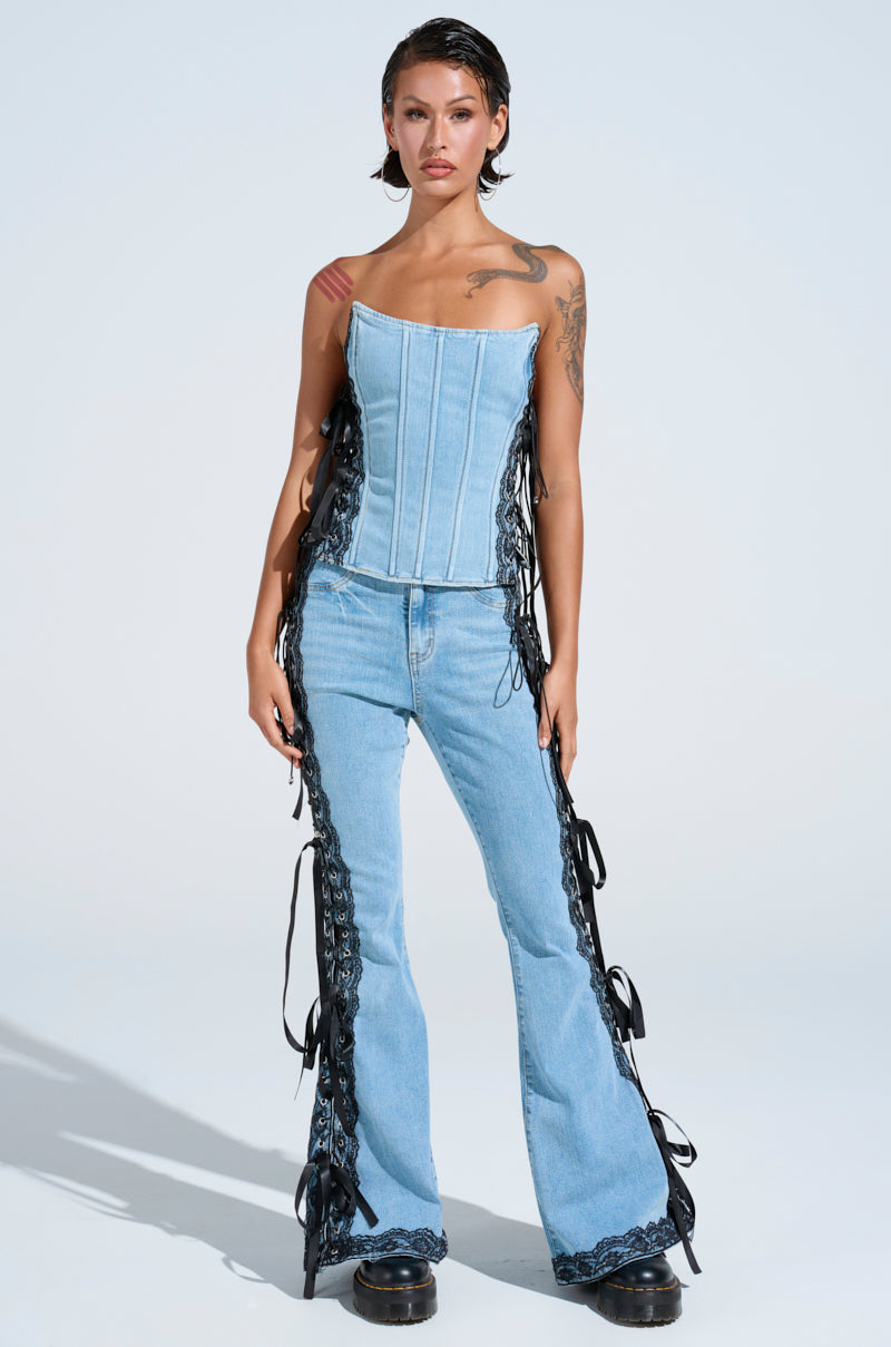 A person poses in light blue denim corset top and TICKED AND TIED LACE AND BOW DETAIL DENIM PANTS with lace-up panels, flared legs. They have short hair, visible tattoos, and black shoes against a plain light-colored backdrop.