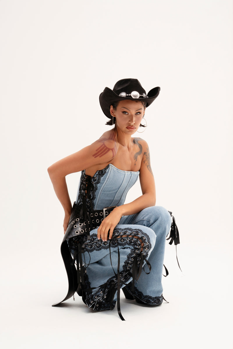 Wearing TICKED AND TIED LACE AND BOW DETAIL DENIM PANTS with decorative black accents and a black cowboy hat, someone confidently poses in a squat against a plain backdrop, highlighting the stylish lace-up panels.