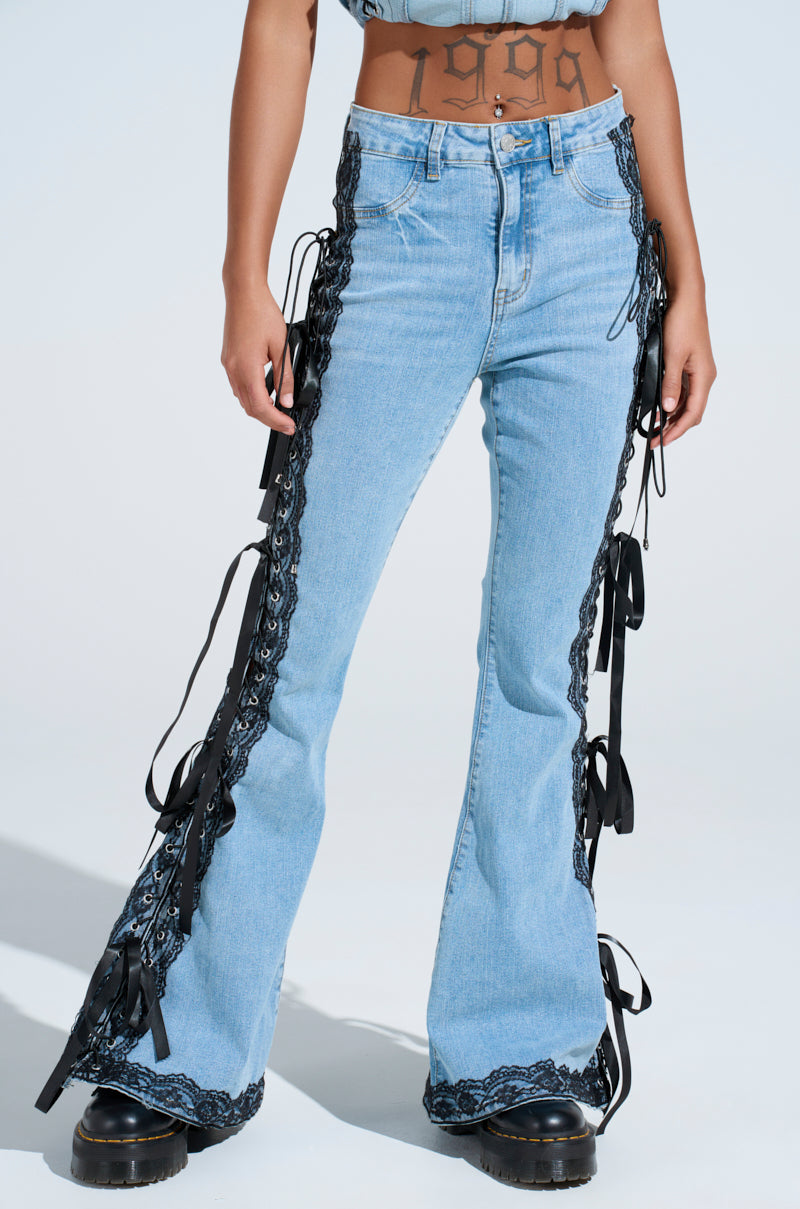 Wearing TICKED AND TIED LACE AND BOW DETAIL DENIM PANTS, a person showcases light blue denim with black lace-up panels and side ribbons. A "1999" tattoo decorates their midriff as they stride in black platform boots. The flared legs create drama against a white backdrop.
