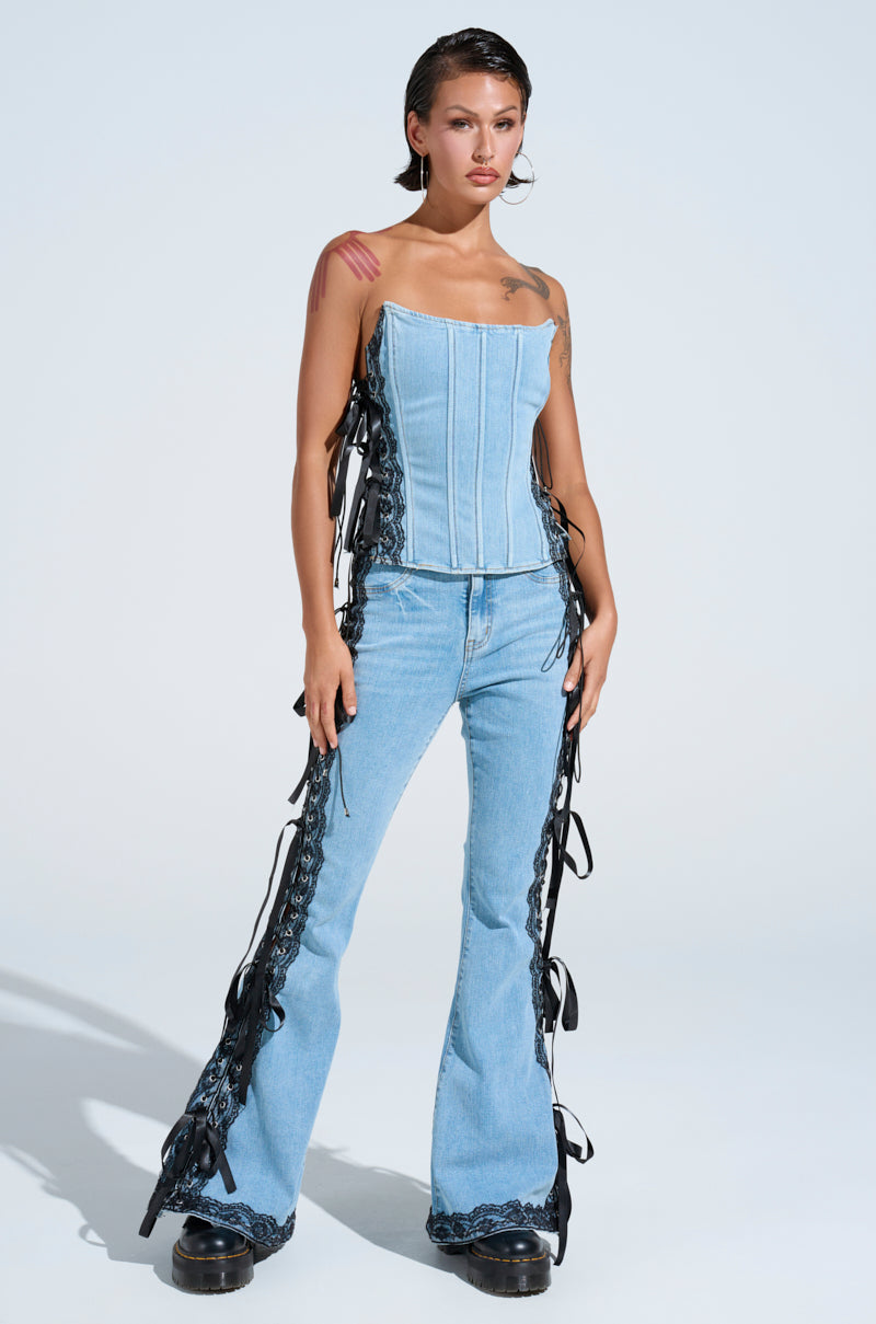 With short hair, large hoop earrings, and black platform shoes, a person exudes effortless style in TICKED AND TIED LACE AND BOW DETAIL DENIM PANTS. These light blue denim pants have a flared leg silhouette and striking black lace-up panels on the sides.