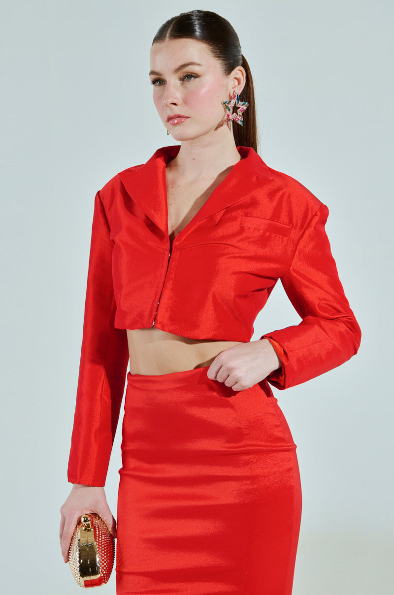 A woman poses against a plain background in a shiny red two-piece outfit featuring a cropped boxy silhouette with a long-sleeved top and high-waisted skirt. Her hair is pulled back, she holds a small gold clutch, and wears star-shaped earrings that catch the light. The ensemble exudes elegance.
