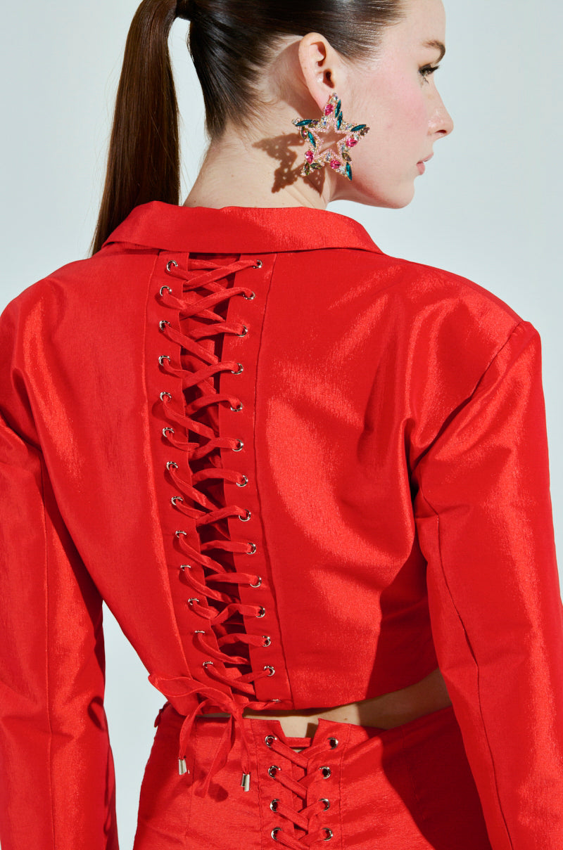 Seen from behind, a person with long hair in a ponytail dons the BRIGHTEST LIGHT BLAZER—a shiny red outfit with a cropped boxy silhouette and corset-style lacing up the back. Star-shaped earrings complement the ensemble, which also features lacing on the lower back.