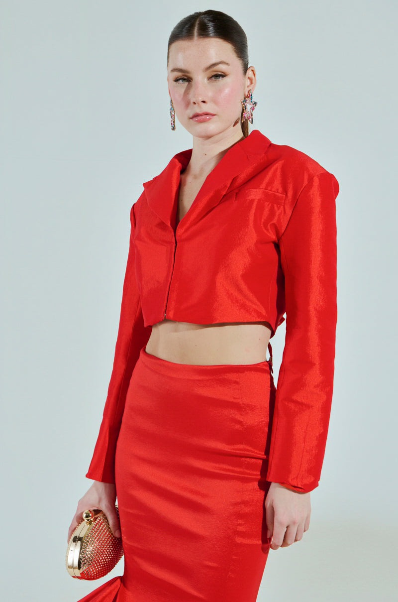 Someone sports the gleaming red BRIGHTEST LIGHT BLAZER with a cropped boxy silhouette, paired with a matching high-waisted pencil skirt and holds a small gold clutch. Their long hair is tied back, complemented by bold red lipstick and earrings, enhancing the satin-lined interior against a plain backdrop.