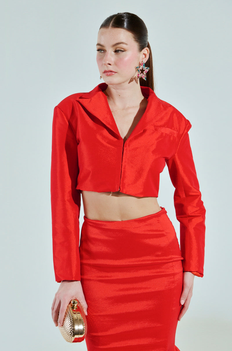 A woman with long brown hair pulled back wears the BRIGHTEST LIGHT BLAZER, a red, long-sleeve crop top with a boxy silhouette, and a high-waisted skirt. She accessorizes with a gold clutch and star-shaped earrings against a plain background.