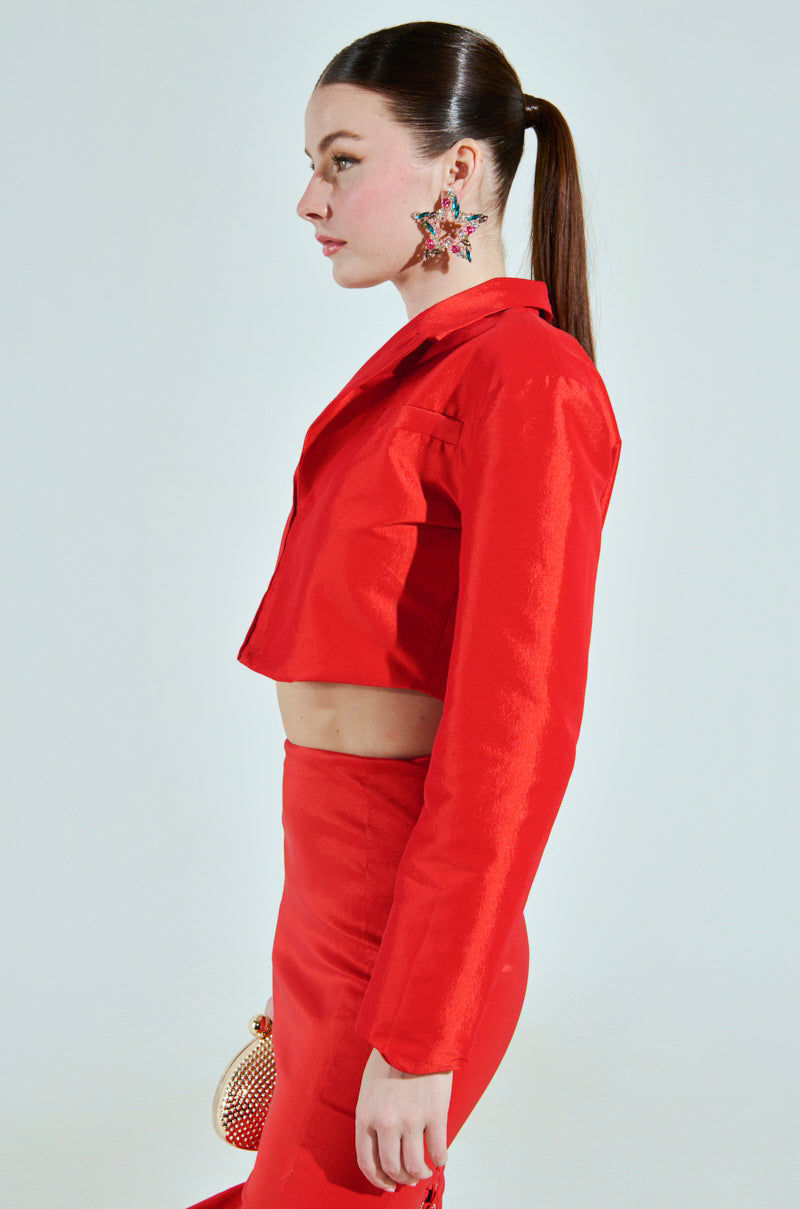 A woman in profile flaunts the BRIGHTEST LIGHT BLAZER, a cropped boxy silhouette with long sleeves, paired with a high-waisted red skirt. Her sleek ponytail and star-shaped earrings highlight the look, completed by a sparkling red purse and satin-lined interior against a plain white backdrop.