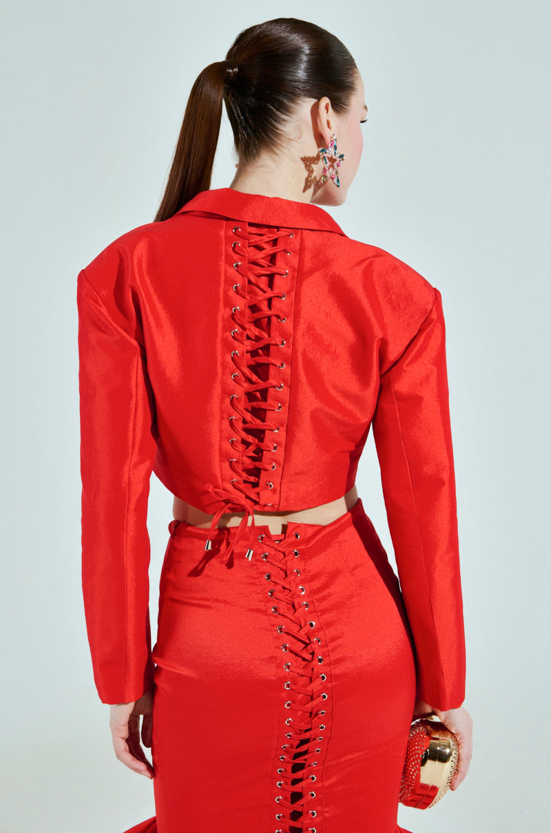 A woman showcases the BRIGHTEST LIGHT BLAZER with lace-up back details and a cropped boxy silhouette. She wears star-shaped earrings, holds a gold handbag, and has a sleek ponytail against a light gray background.