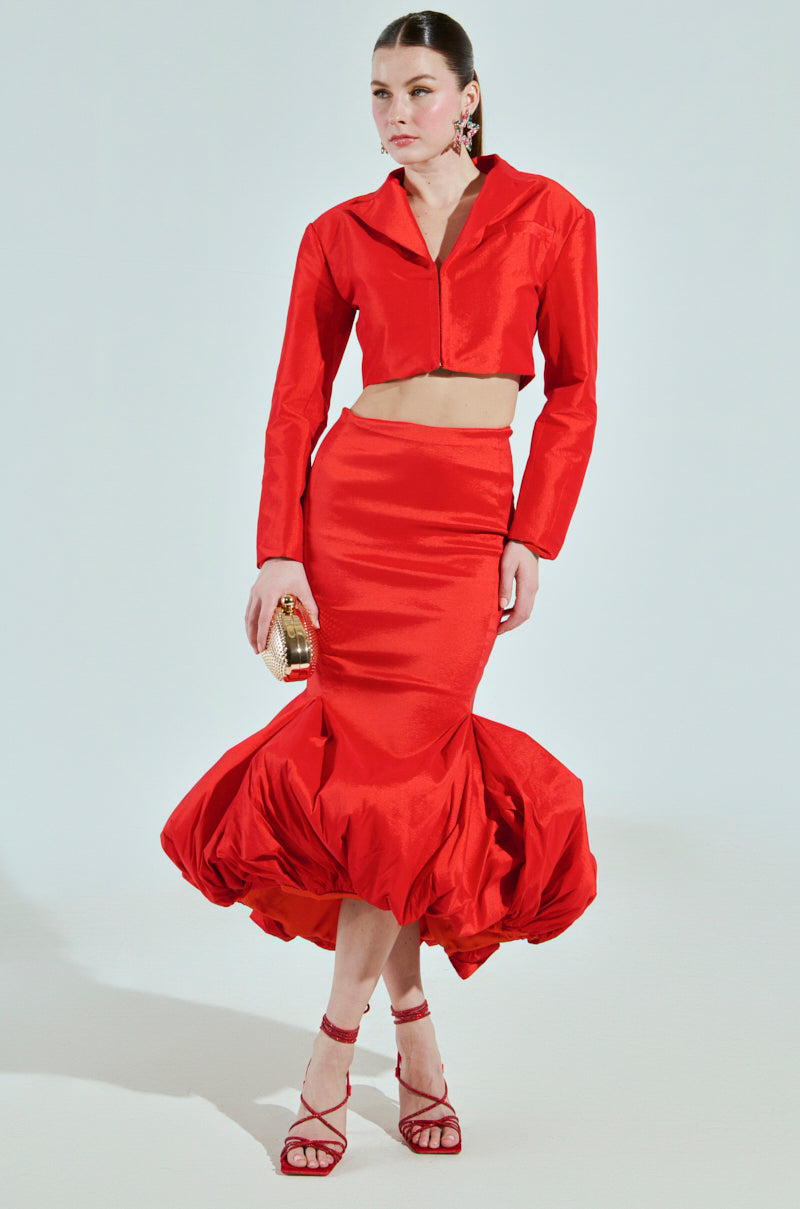 A model in a striking red ensemble, featuring the BRIGHTEST LIGHT BLAZER, stands against a plain background. She showcases a collared, long-sleeve crop top and high-waisted skirt with red strappy heels and a small clutch.